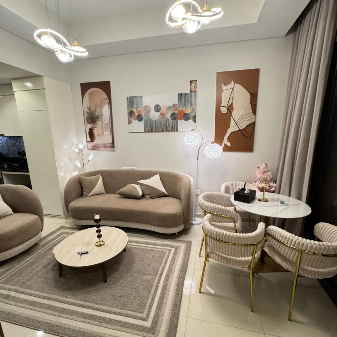 Living room with beige sofa, round marble table, and four chairs with gold legs. Artwork adorns the walls.