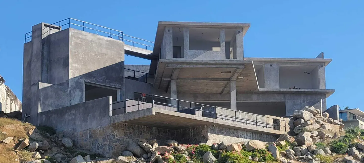 Unfinished gray concrete house with black railings on a rocky hillside against a clear blue sky.
