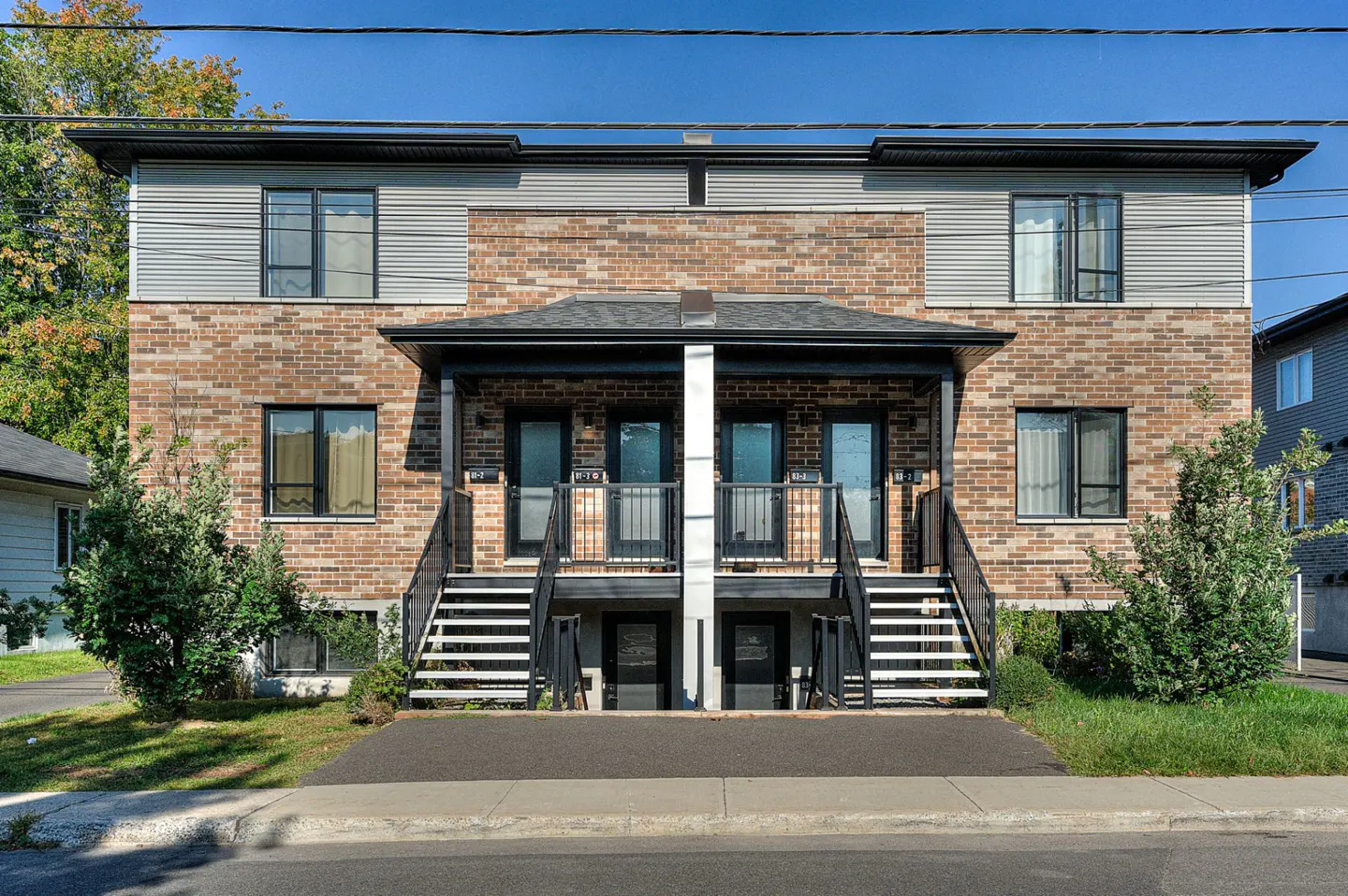 Two-story duplex with brick and gray siding, black doors, and staircases leading to the entrances. Green trees and grass surround the building.