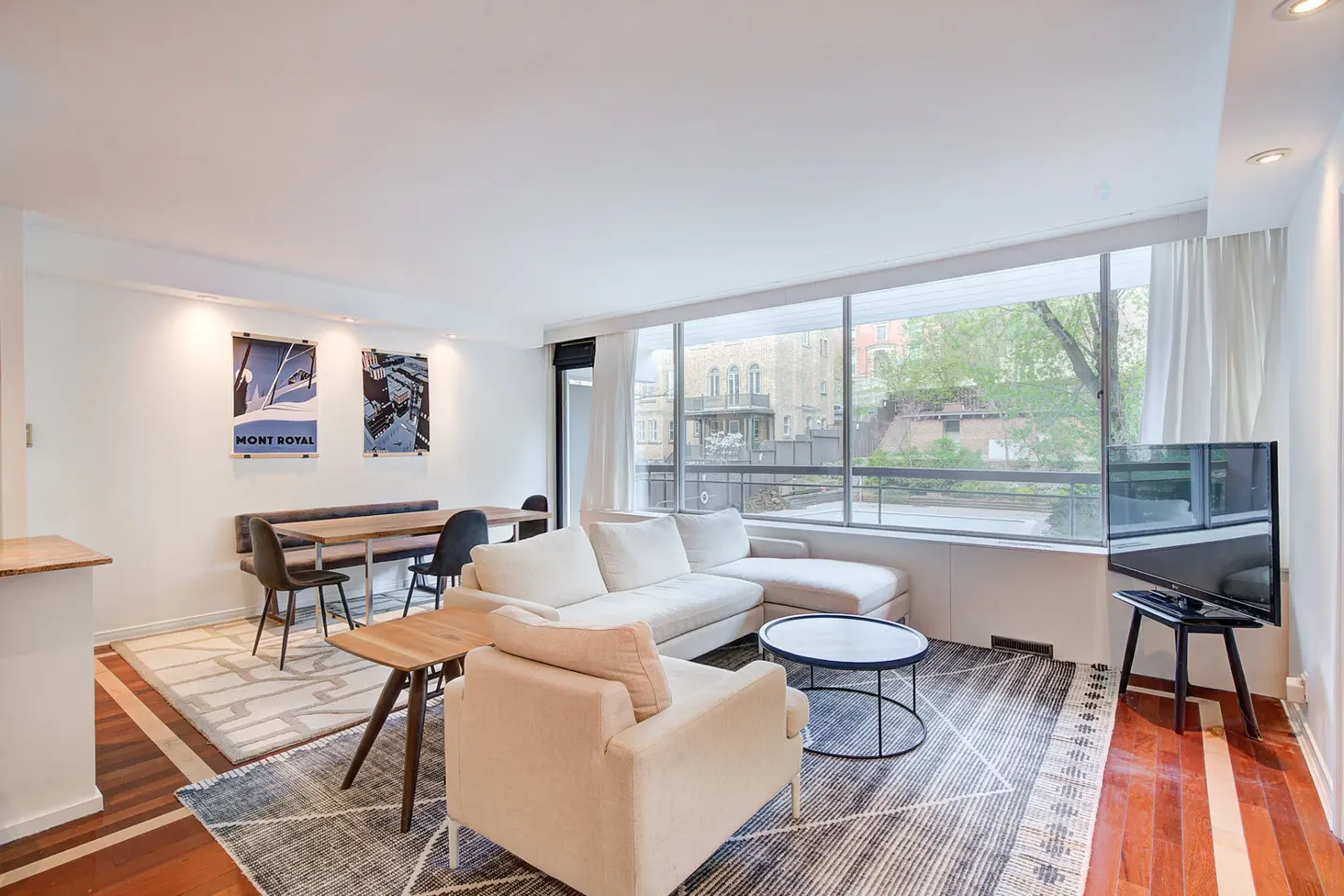 Bright living room with white walls, wood floors, and large windows. Beige sectional sofa, armchair, and patterned rug. Dining table with chairs and wall art.