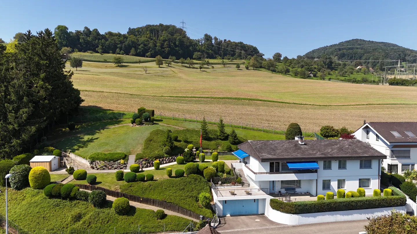 A white two-story house with blue awnings sits on a hill with a manicured lawn and a field in the background.