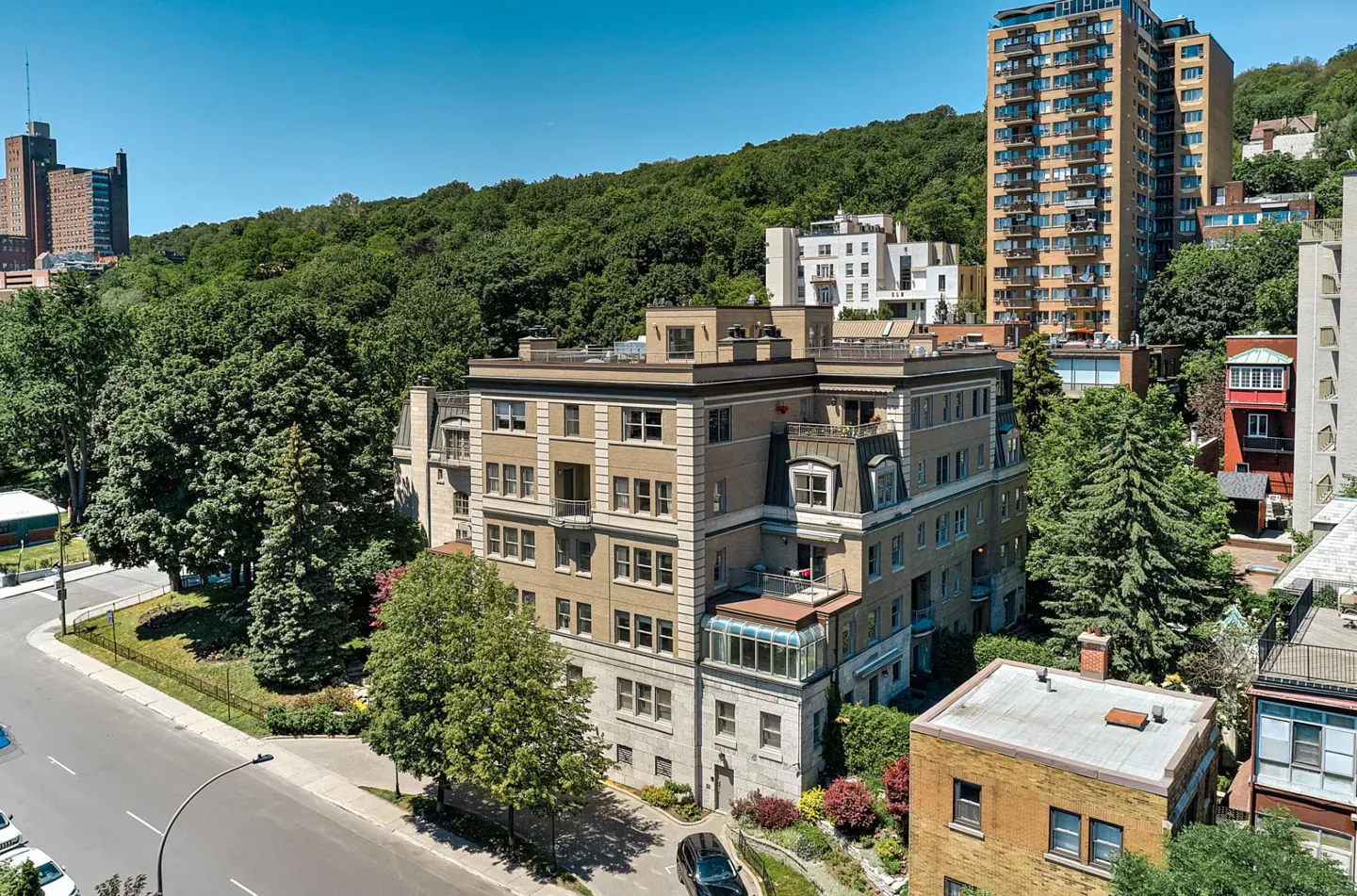 Aerial view of a beige apartment building with a glass sunroom, surrounded by lush green trees and other buildings.