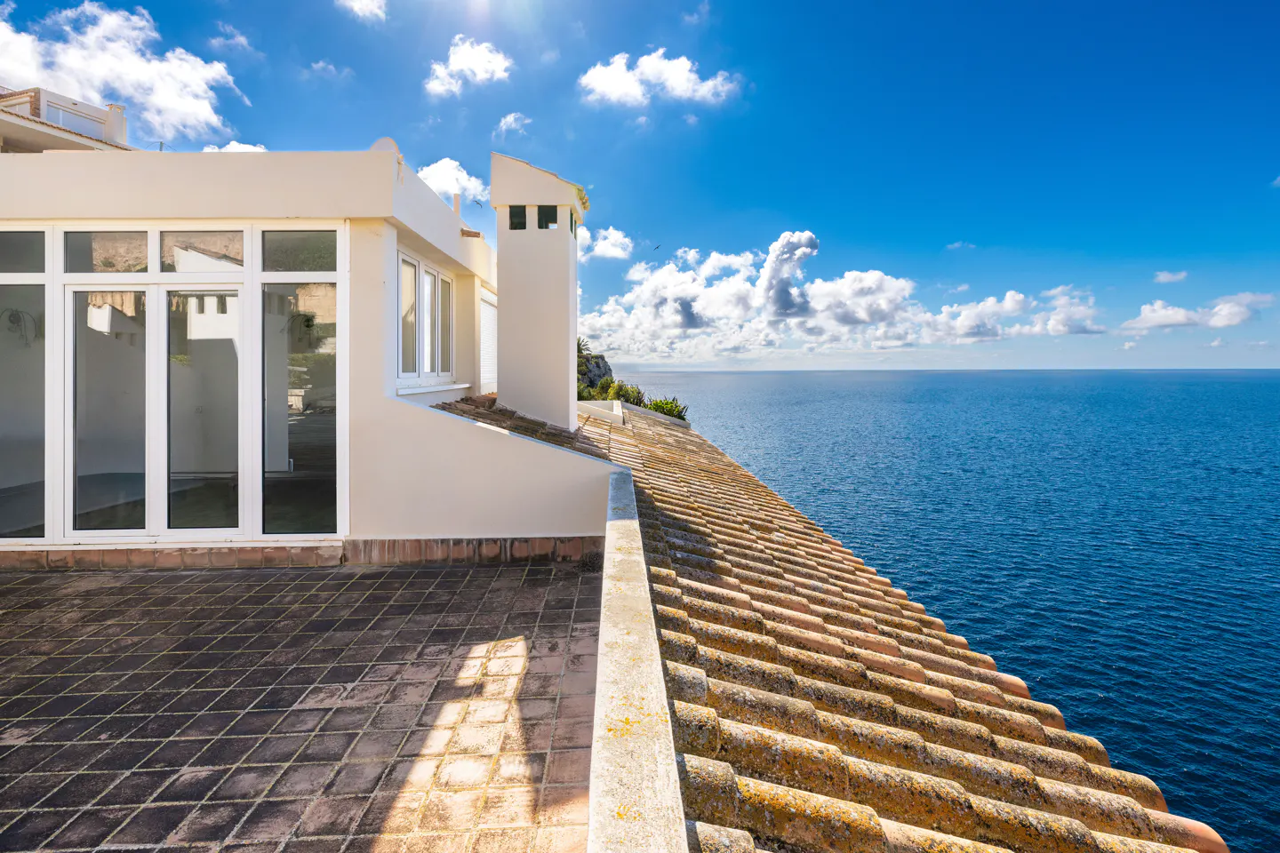 A cream-colored building with a tiled roof overlooks the blue ocean under a sunny, cloudy sky.