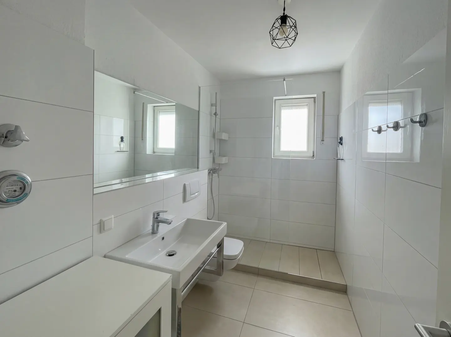 Bright, modern bathroom with white tiled walls, a large mirror, and a white sink with chrome fixtures. A geometric light fixture hangs from the ceiling.