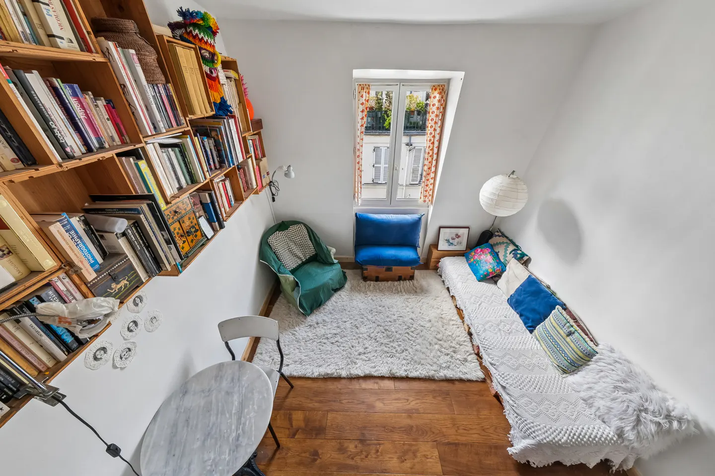 Cozy attic room with a sloped bookshelf, green chair, blue seat, and a daybed with pillows. A white rug covers the wood floor.