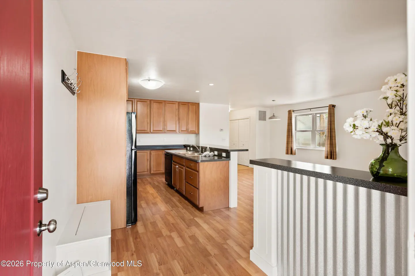 A view of a kitchen and living room with wood floors, cabinets, and a black refrigerator. A red door is on the left.