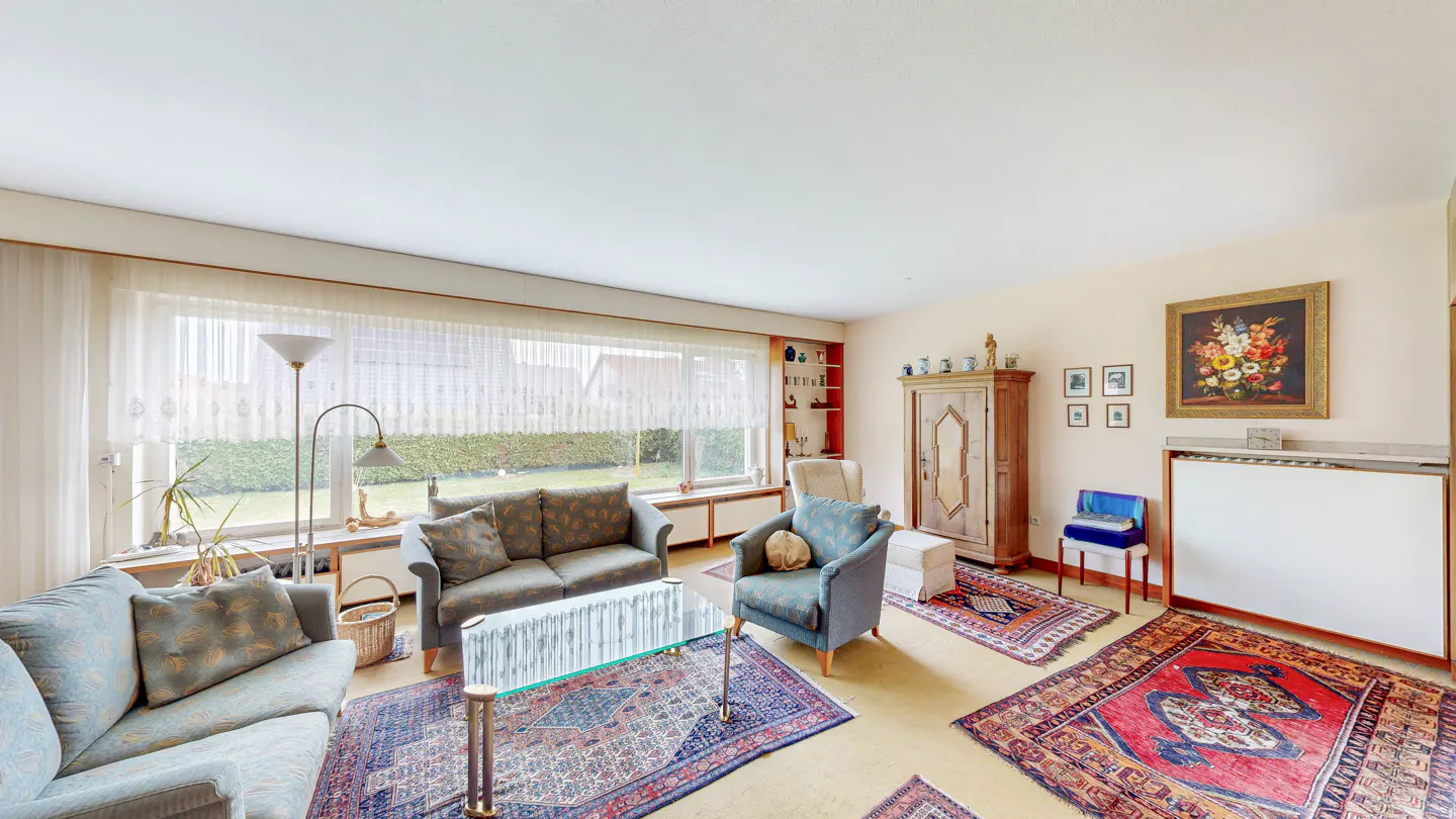 Bright living room with sofas, armchairs, and Persian rugs on a yellow floor. A large window overlooks a green lawn.