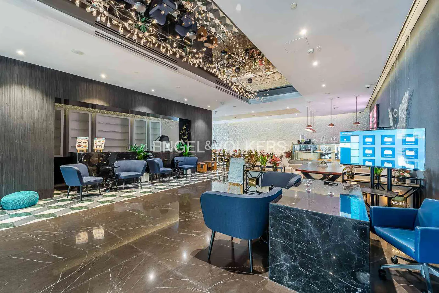 Engel & Völkers office lobby with blue chairs, marble floors, and a mirrored ceiling with decorative lights. A large screen displays files.