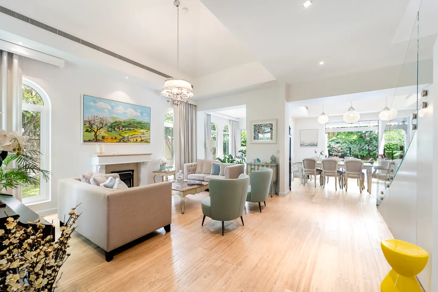 Bright, open-plan living and dining area with light wood floors, white walls, and neutral furniture. A landscape painting hangs above a fireplace.