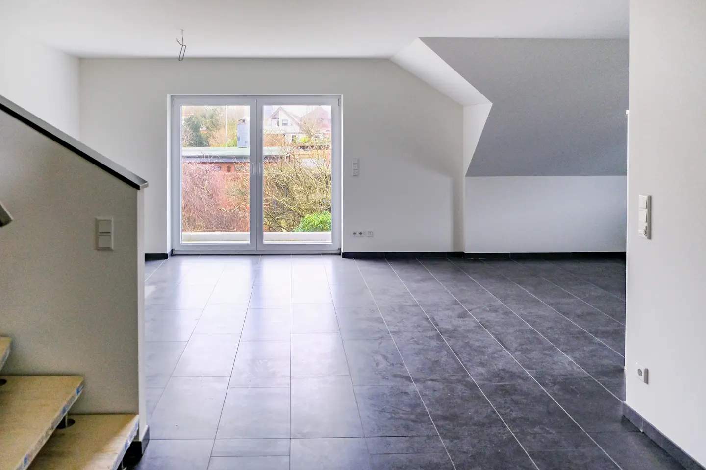Bright, empty room with dark tile floor, white walls, and a glass door to the outside. Stairs are visible on the left.