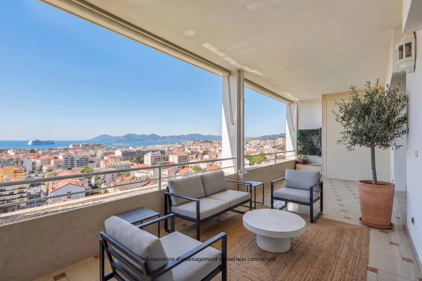 Balcony with gray seating overlooks a city and ocean view. A round white table sits on a woven rug. A potted tree is on the right.