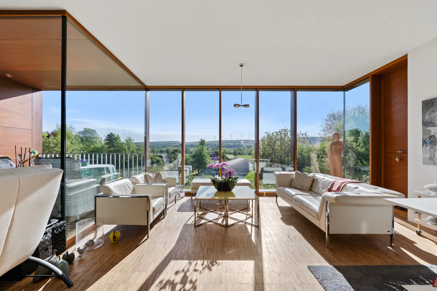 Bright living room with floor-to-ceiling windows showing a green landscape. White sofas and chairs sit on a wood floor.