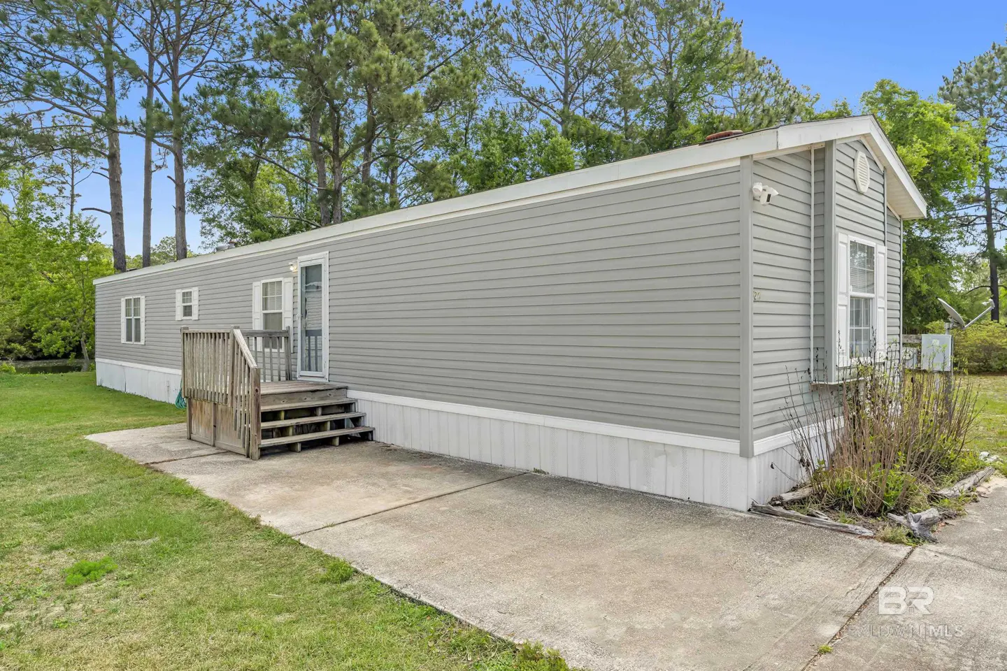Exterior view of a gray mobile home with a wooden porch and concrete driveway, surrounded by green grass and trees.