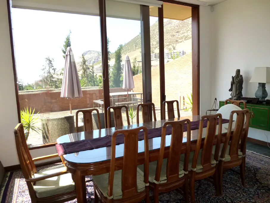 Bright dining room with a long wooden table and chairs, a purple runner, and a view of a patio with umbrellas and a mountain.