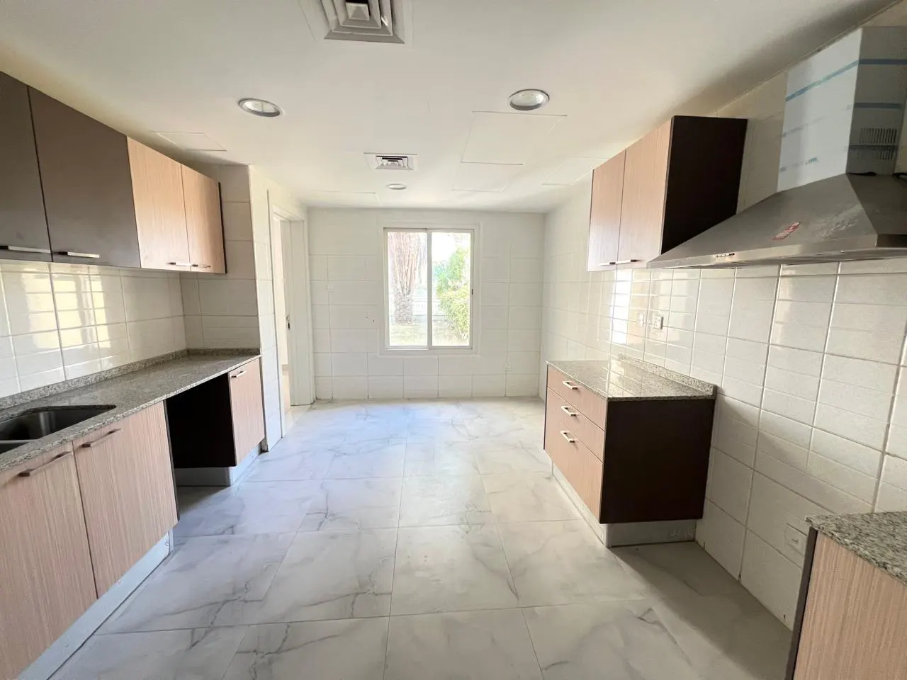A bright kitchen with white tiled walls and marble floors. Brown and tan cabinets line the walls, with a window in the center.