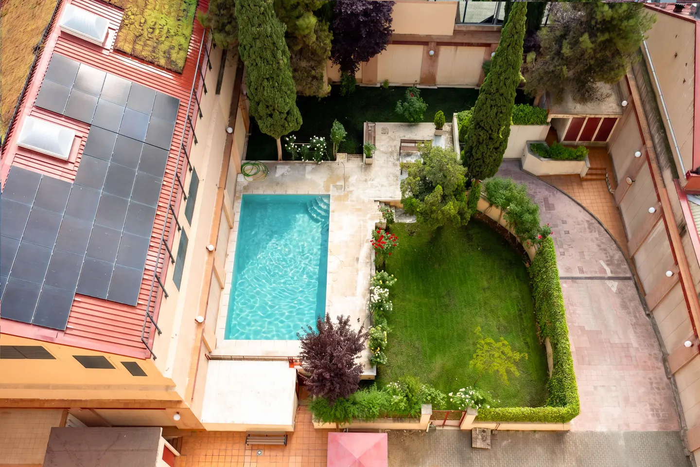 Aerial view of a building with solar panels, a pool with turquoise water, and a green lawn with trees and flowers.
