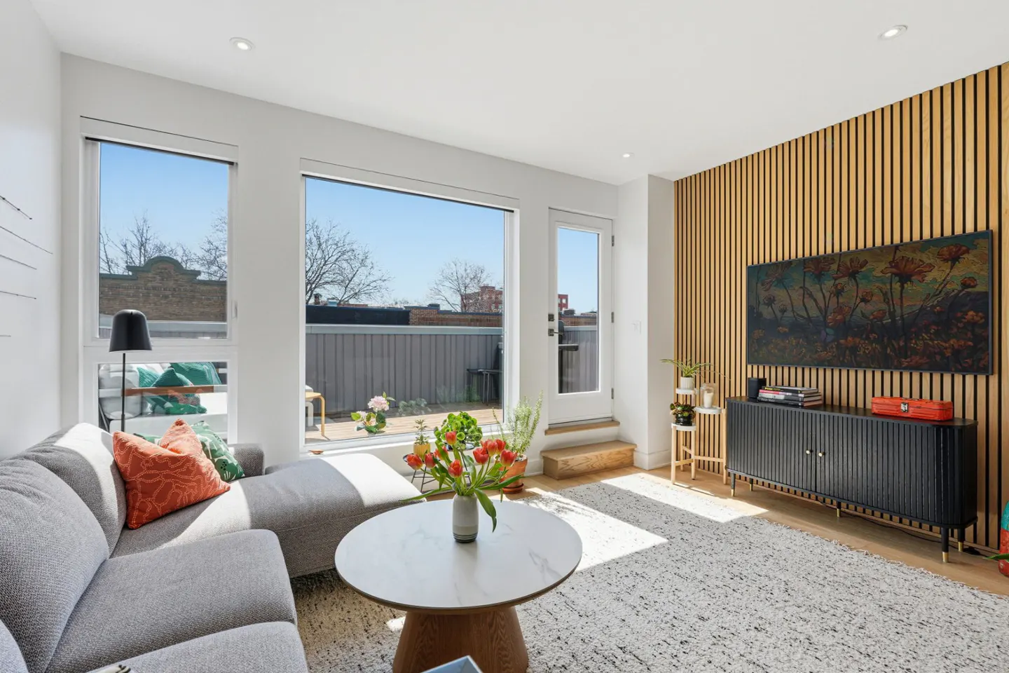 Bright living room with gray sectional, round table with tulips, and black cabinet under a floral art piece on a wood slat wall.