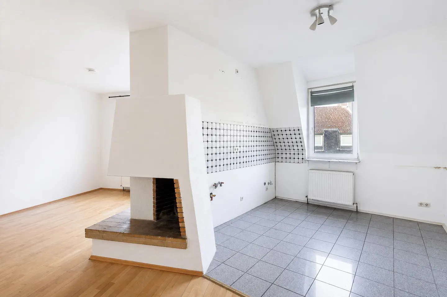 Bright, empty room with a fireplace, hardwood floors, and gray tiled kitchen area with black and white backsplash. A window is visible.