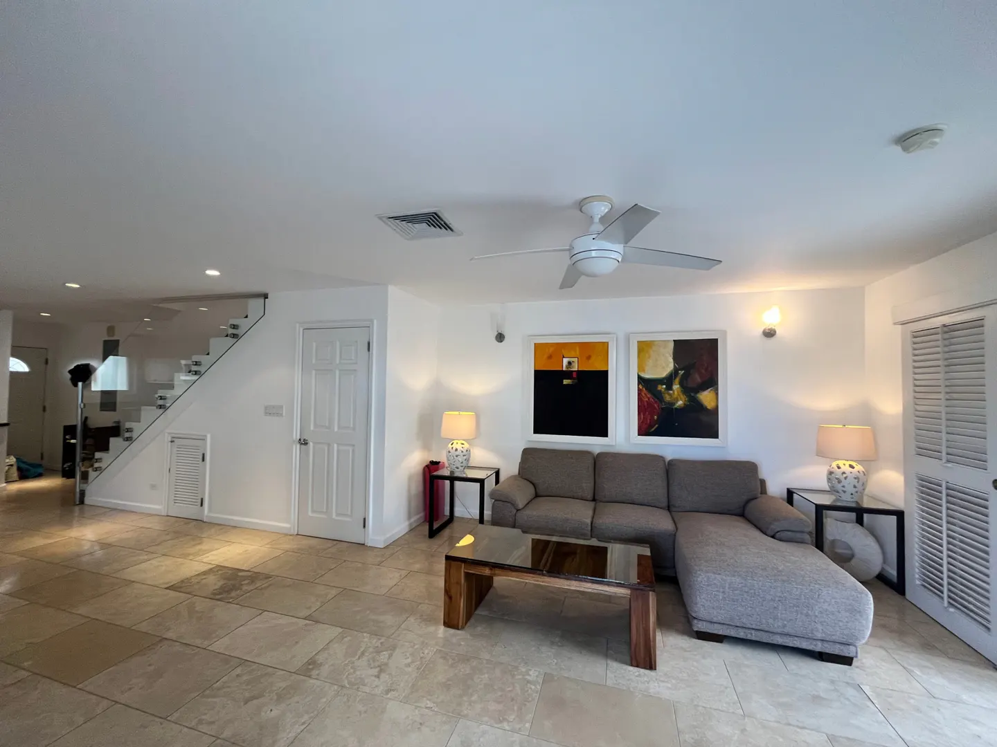 Living room with tile floors, a gray sectional sofa, and a wooden coffee table. Two paintings hang above the sofa. A staircase is visible in the background.