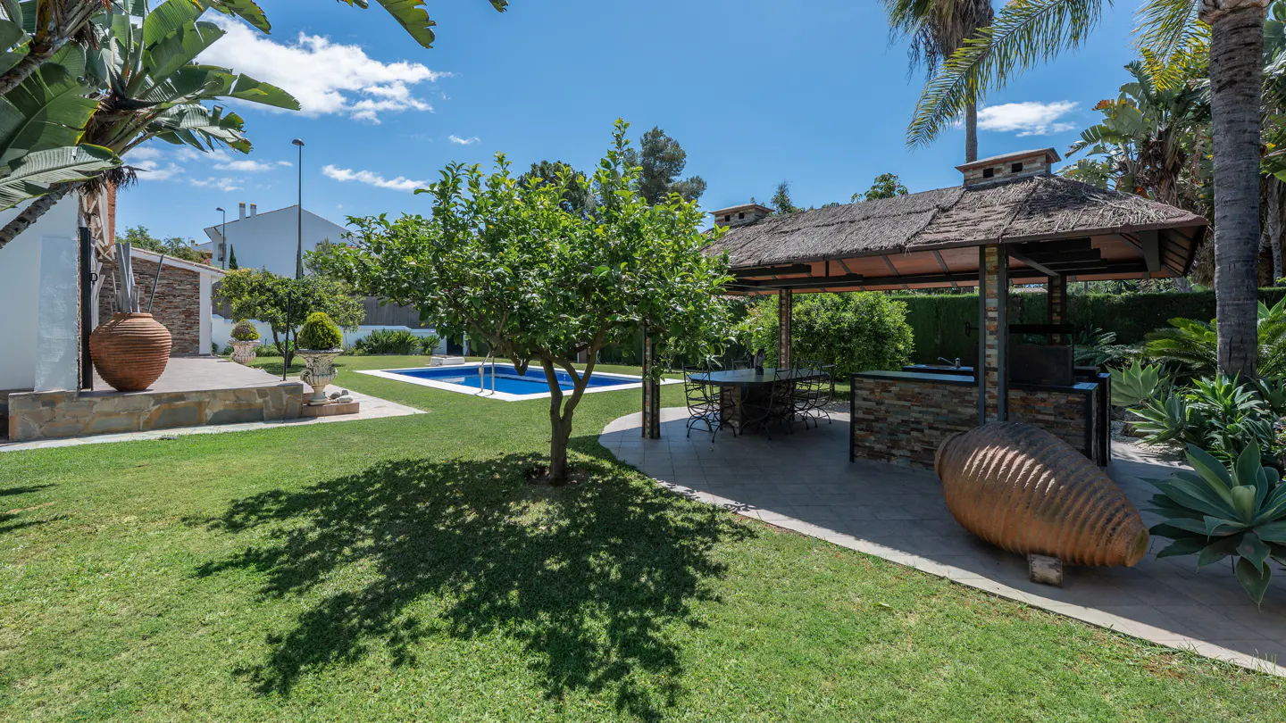 Lush green backyard with a pool, thatched-roof gazebo, and large terracotta pots on a sunny day.
