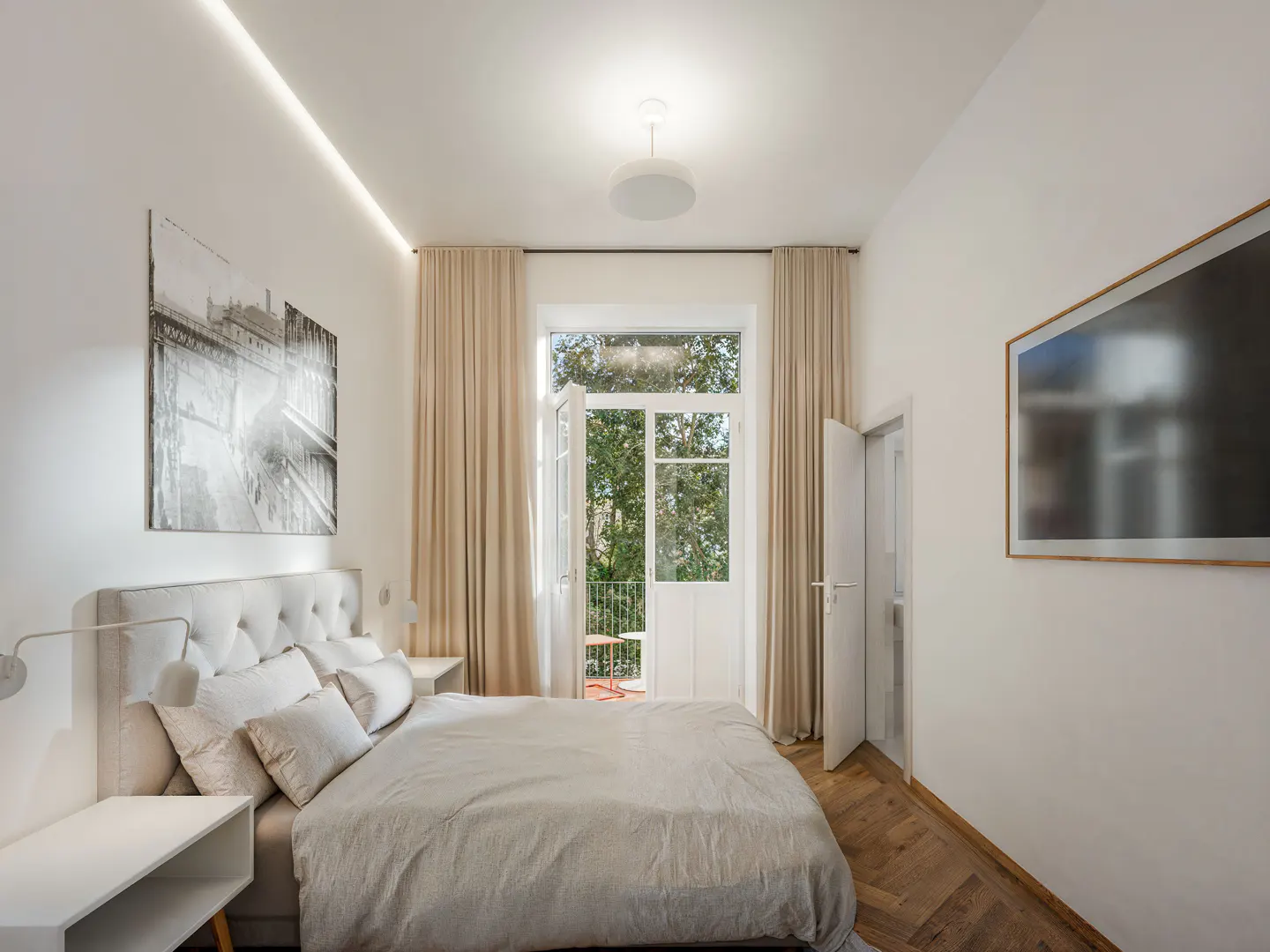 Bedroom with a queen bed, white walls, wood floors, and a balcony with open doors. A black and white picture hangs above the bed.