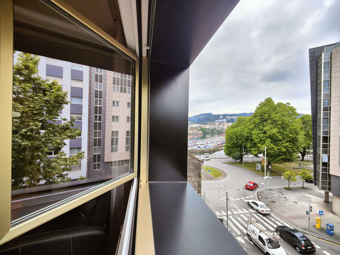 View from a modern apartment window, showing city buildings, green trees, and traffic on a cloudy day.