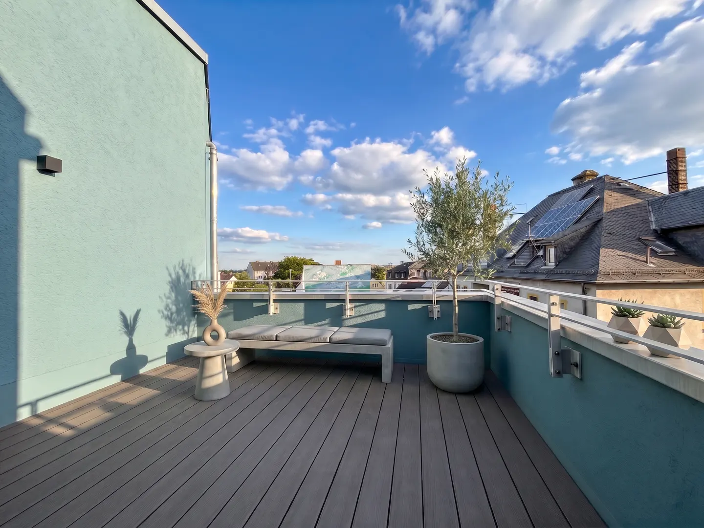 Rooftop deck with gray wood planks, a bench, and a small tree in a pot. Blue sky with clouds in the background.