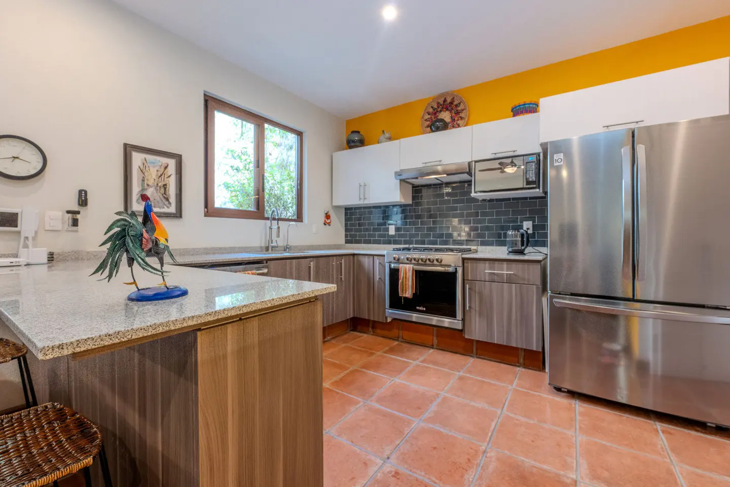 Bright kitchen with terracotta tile floor, stainless steel appliances, and gray cabinets. A yellow accent wall adds a pop of color.