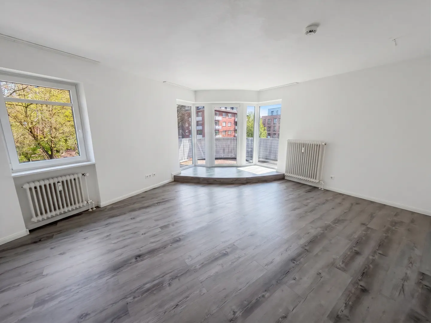 Bright, empty room with gray wood floors, white walls, and a bay window leading to a balcony. A radiator sits under a window with a tree view.