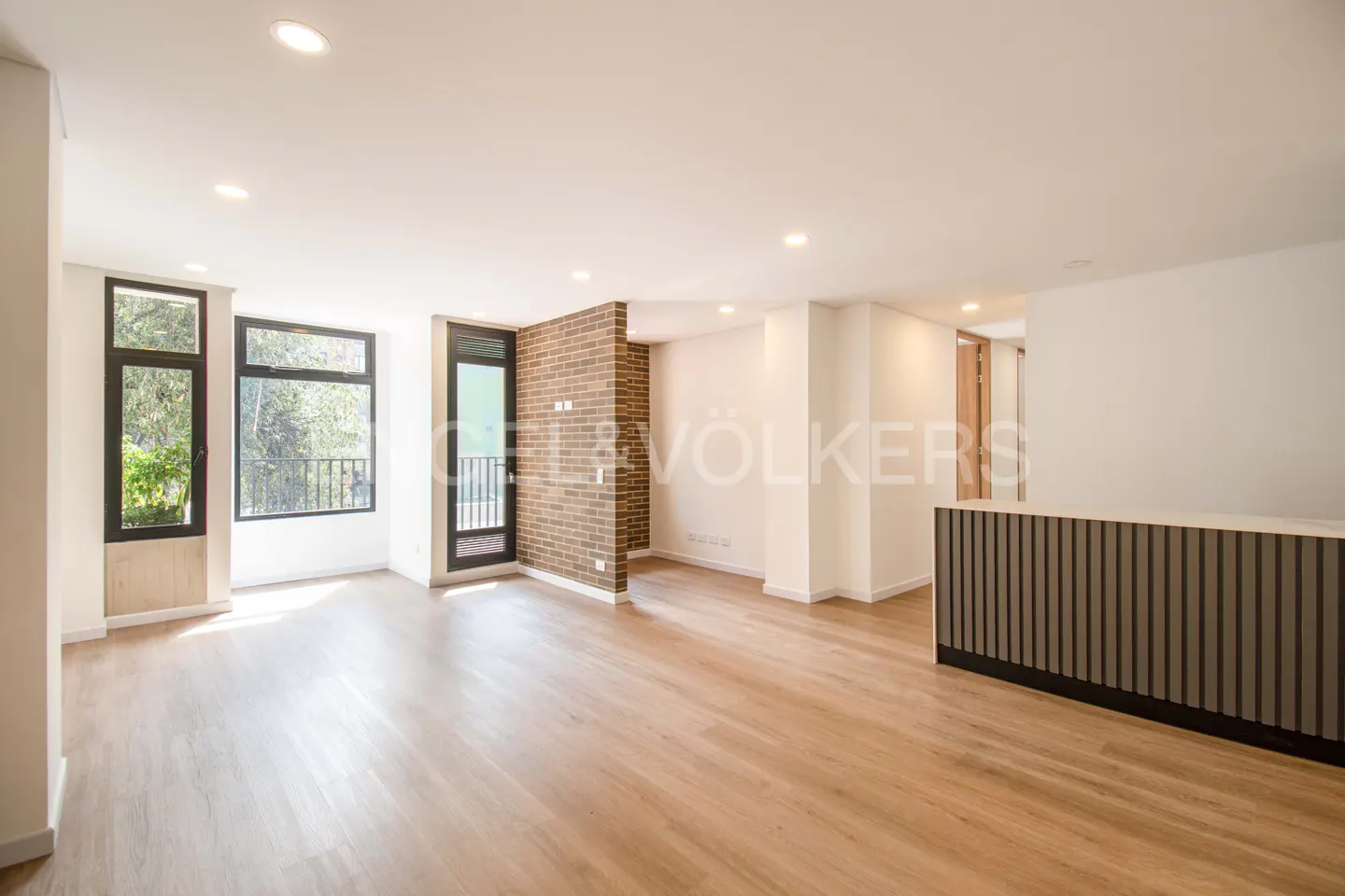 Bright, empty living room with wood floors, white walls, and large windows. A brick accent wall and a dark gray counter add contrast.