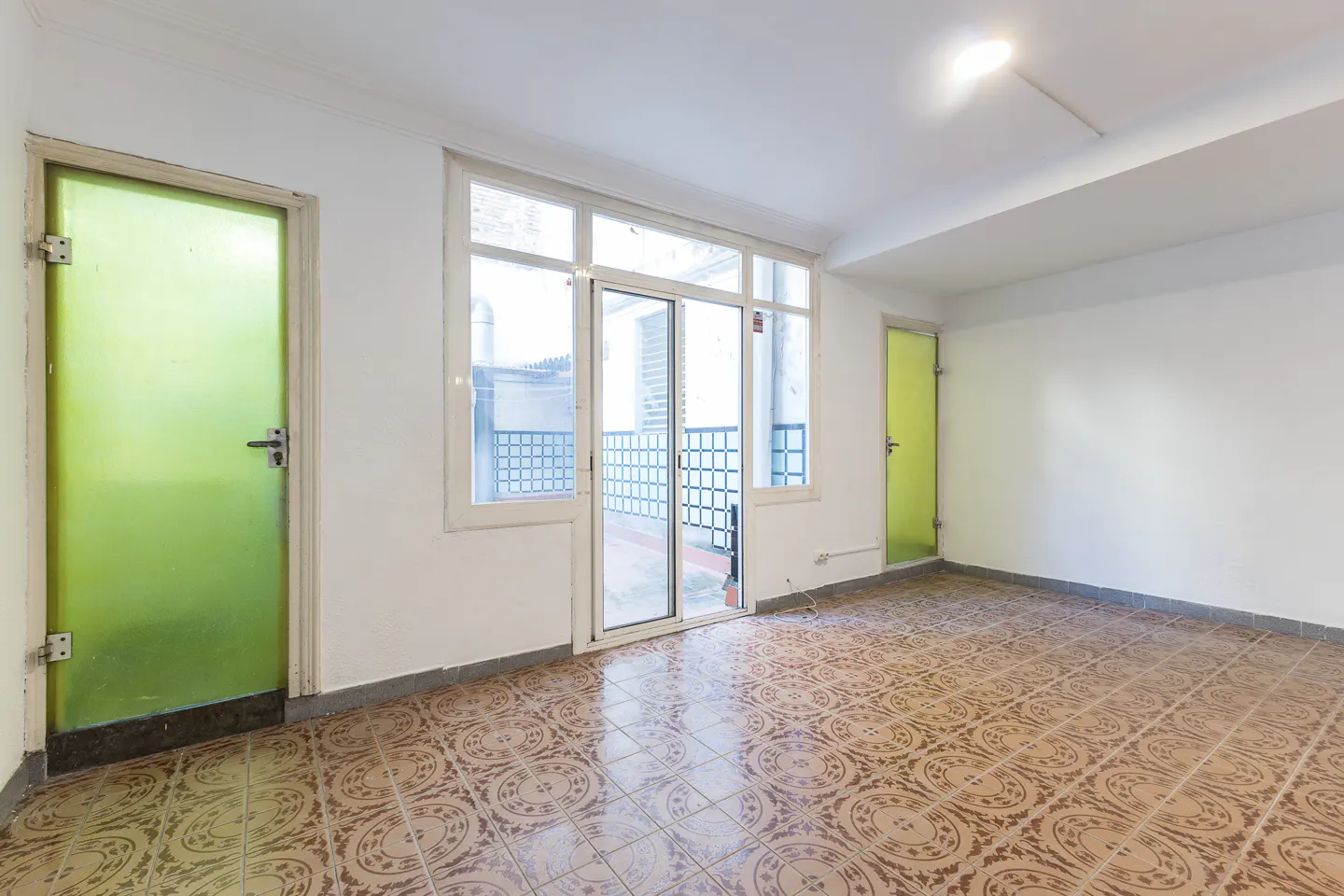 Bright, empty room with patterned tile floor, white walls, and two green doors. A sliding glass door leads to a balcony.