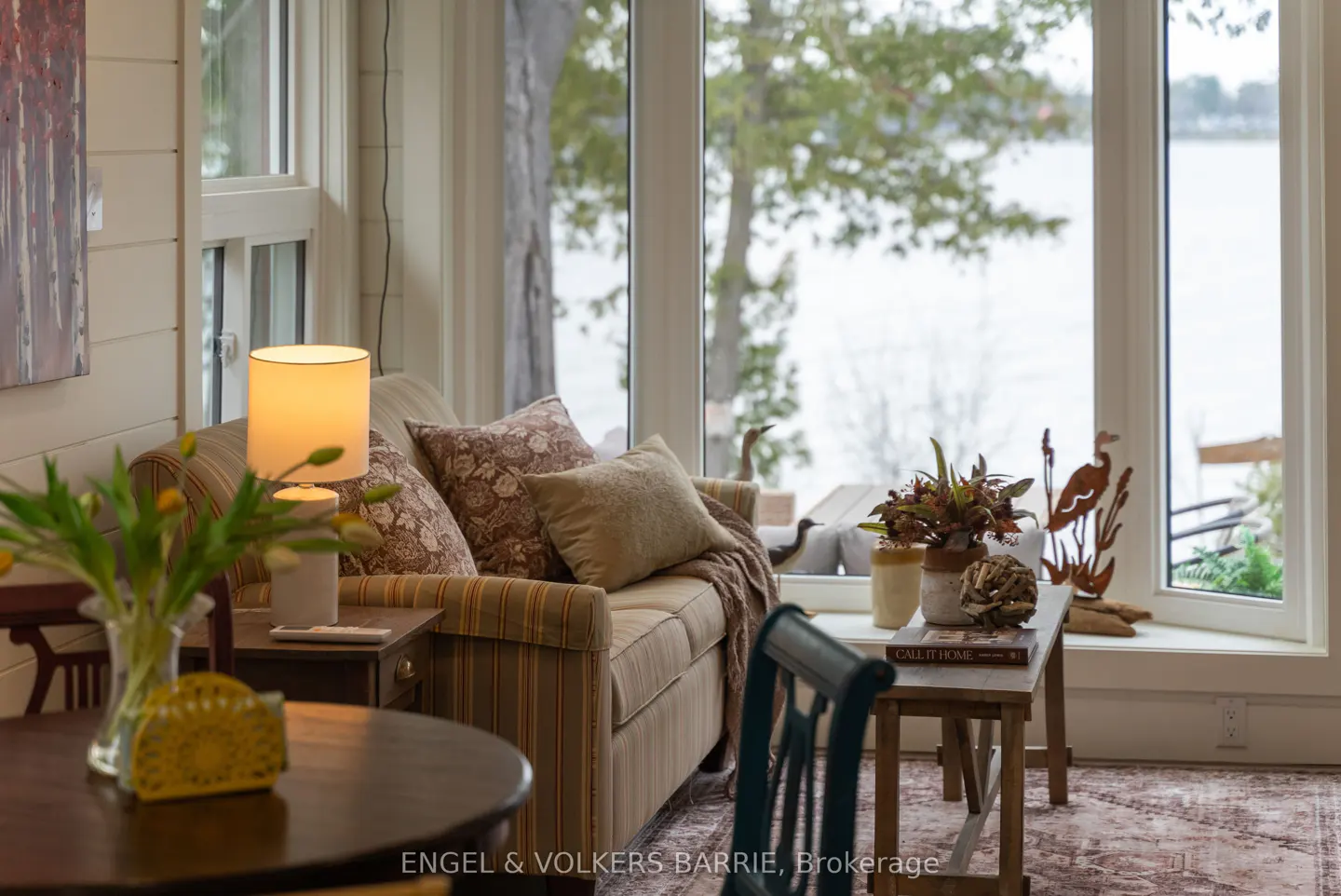 Cozy living room with a striped sofa, floral pillows, and a view of the lake through large windows. A lamp illuminates the scene.