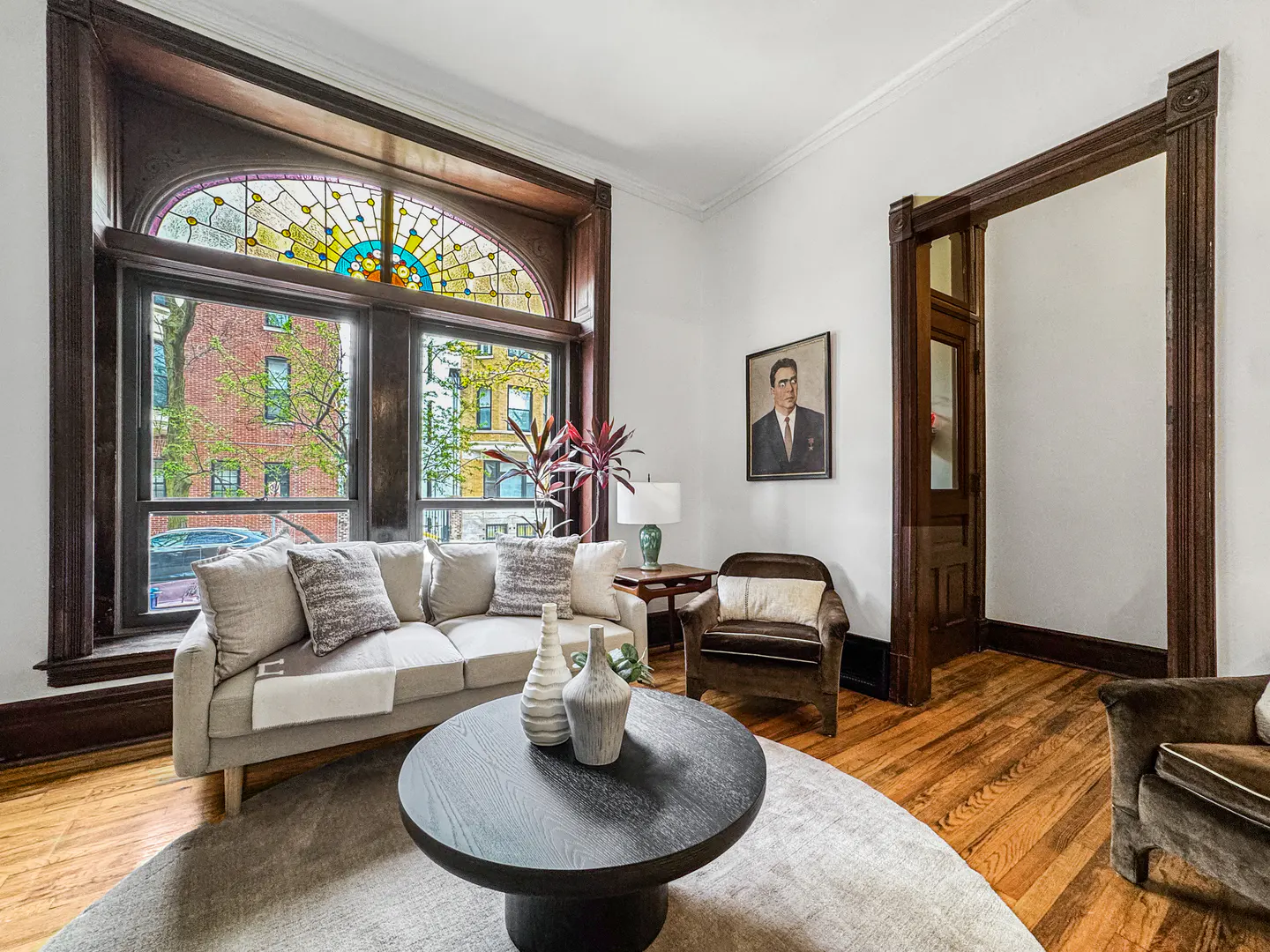 Living room with a beige sofa, round black table, and stained-glass window. A portrait hangs on the white wall.