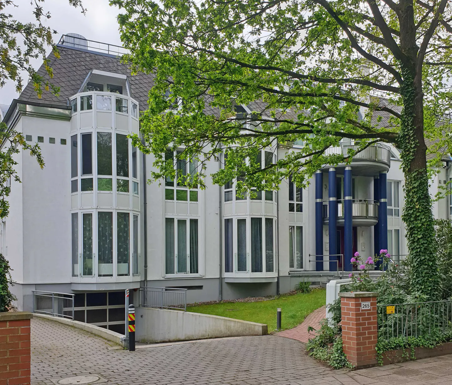 A three-story white apartment building with blue columns and a gray roof. A tree with green leaves is in the foreground.