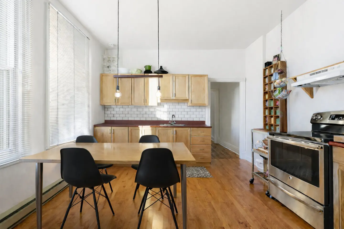 Bright kitchen with wood floors, cabinets, and table. Black chairs surround the table under a modern light fixture. Stainless steel oven on the right.