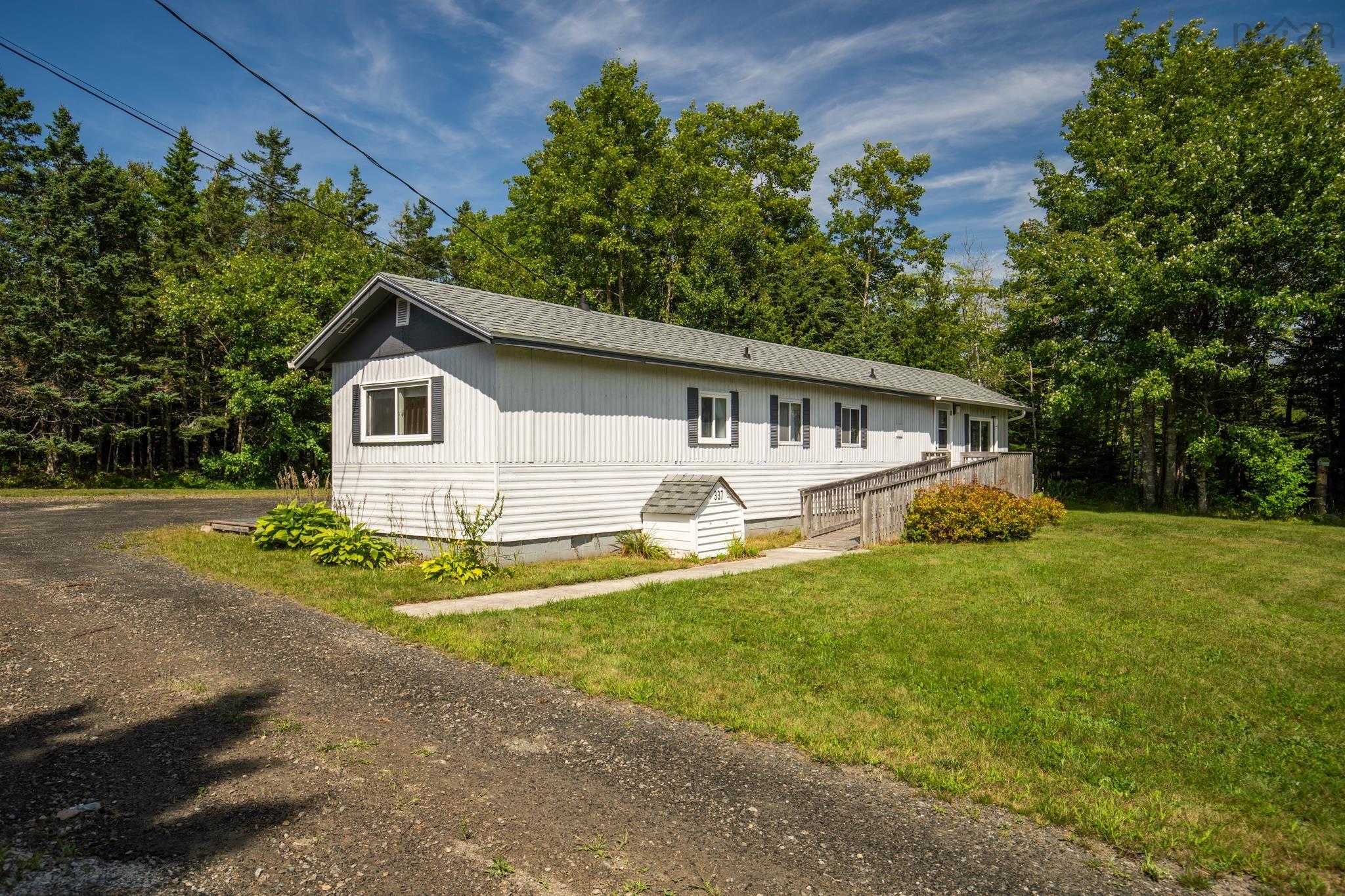 A white single-story home with a wheelchair ramp, gravel driveway, and lush green trees under a blue sky. [AI-generated text]