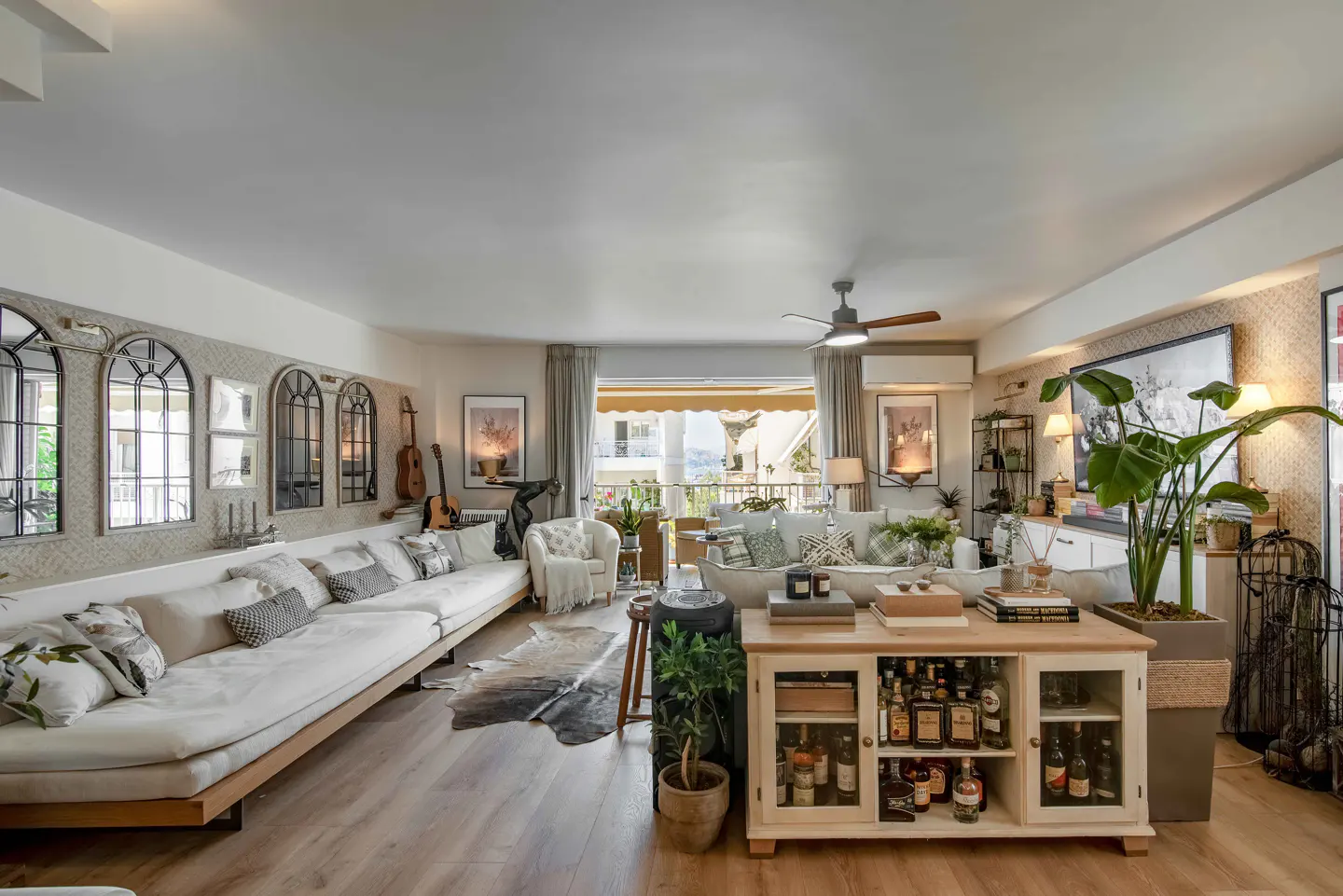 Spacious living room with a long white sofa, a wooden bar cabinet, a large potted plant, and a balcony view.