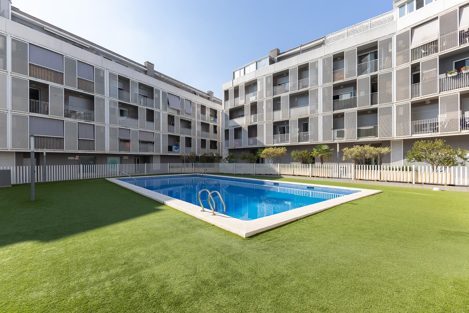 A rectangular swimming pool with blue water sits in a courtyard surrounded by modern apartment buildings and green turf.