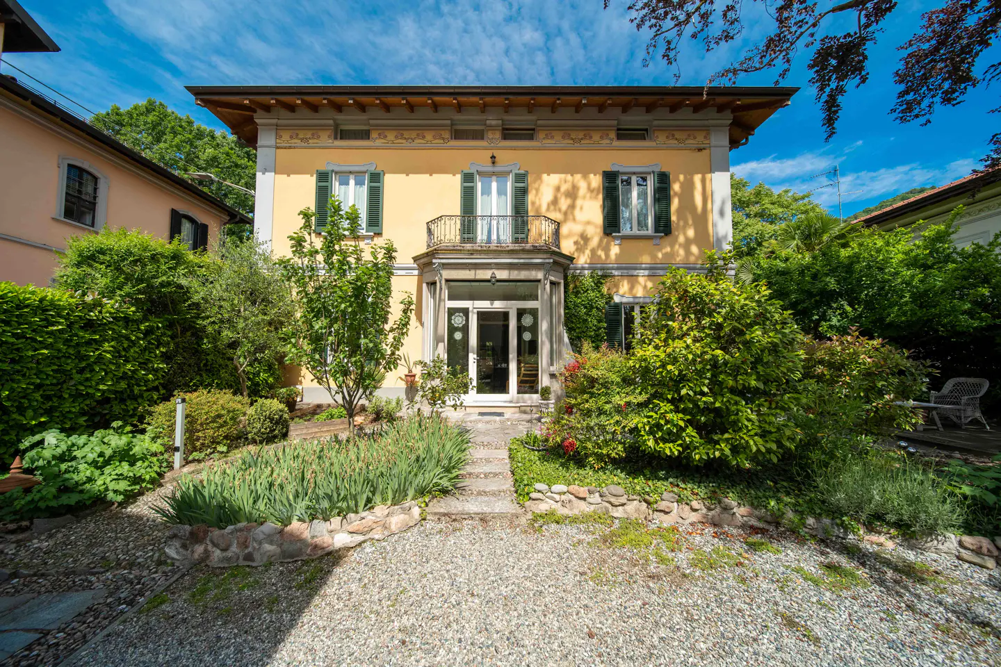Two-story yellow house with green shutters and a small balcony. A stone path leads to the front door, surrounded by lush greenery.