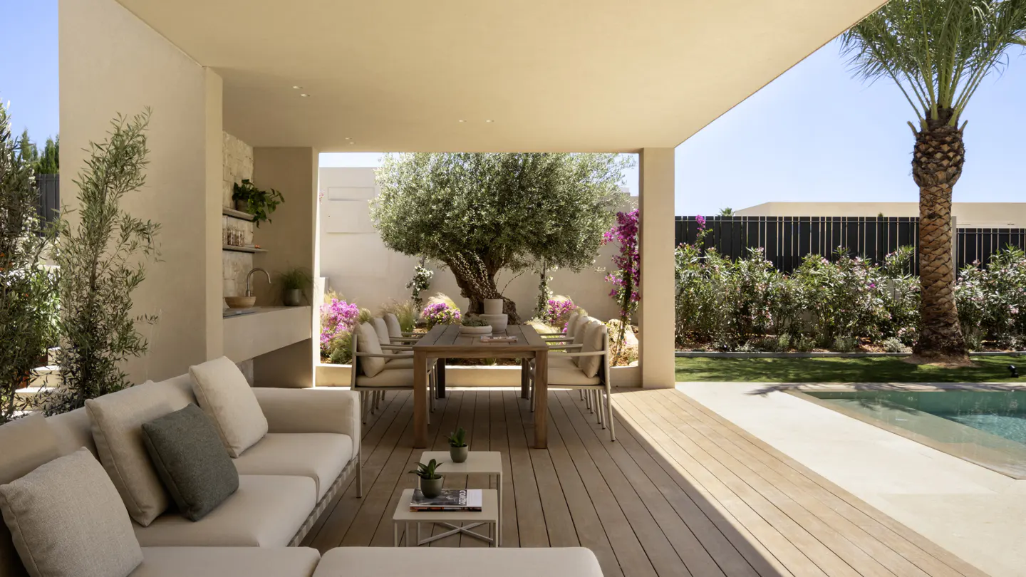 Outdoor patio with a cream sofa, wooden table and chairs, and a tree in the center. A pool and palm tree are visible in the background.