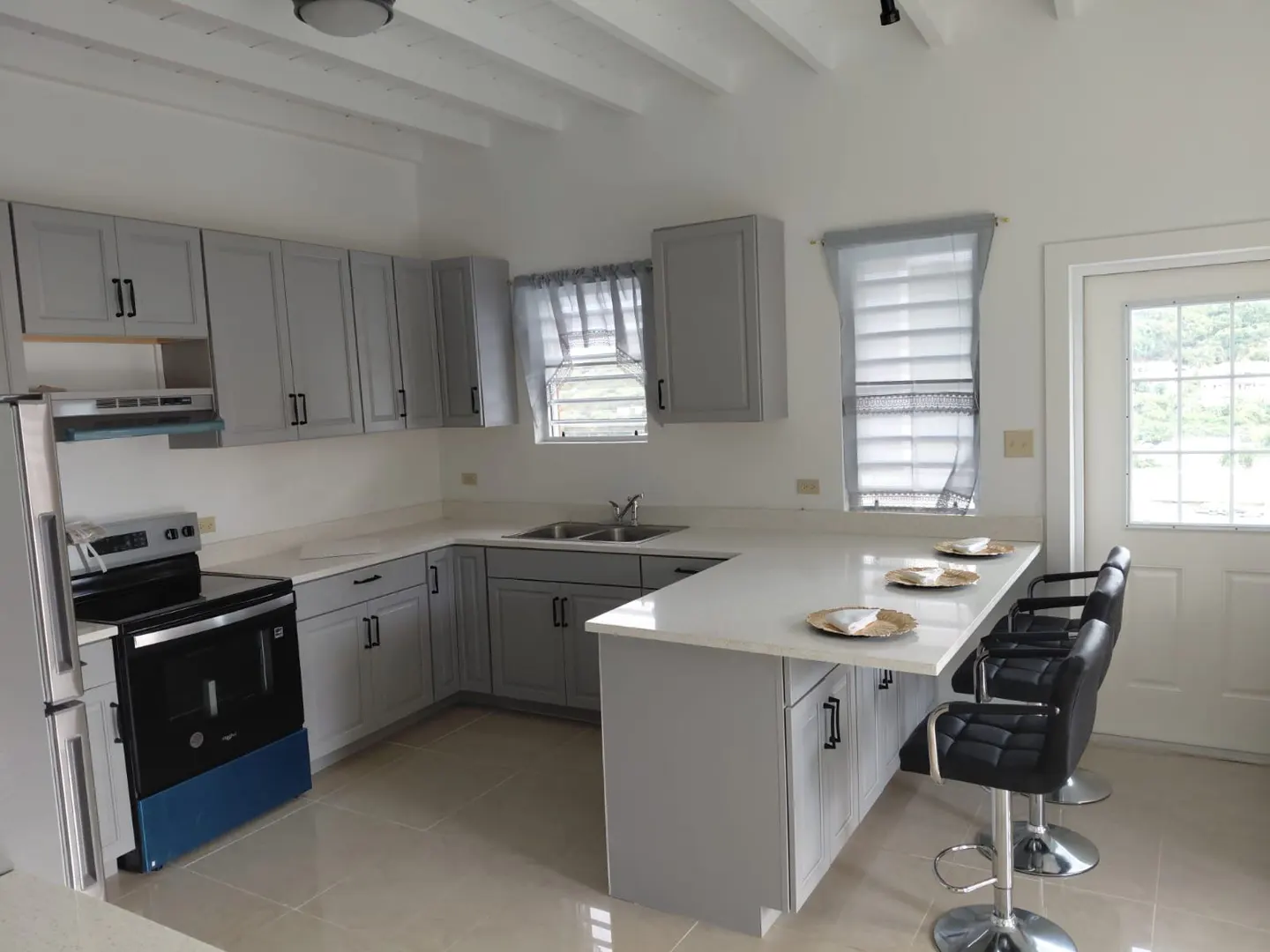 Bright kitchen with gray cabinets, white countertops, and black bar stools at the island. A stainless steel refrigerator and oven are visible.