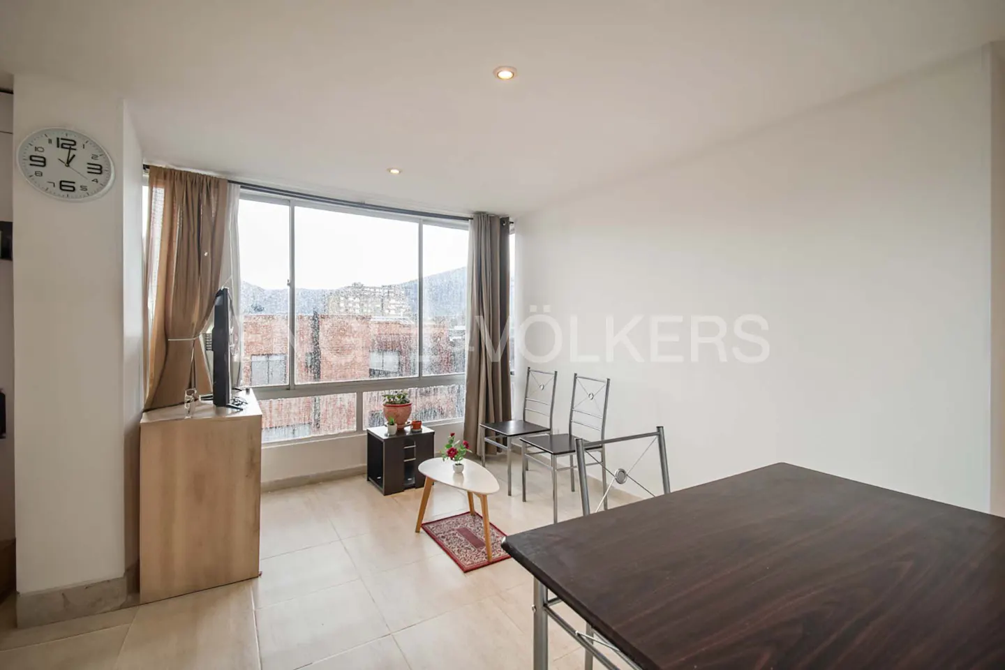 Bright apartment living area with a dark wood table, two metal chairs, a small coffee table, and large windows with curtains.