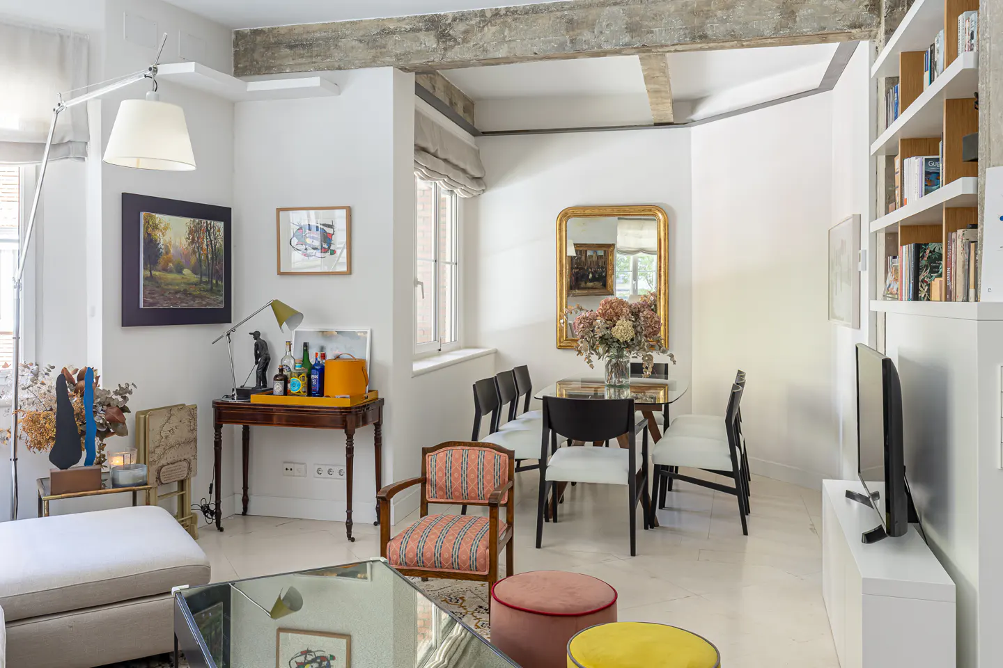 Bright living room with white walls, concrete beams, and a glass dining table with black chairs. Artwork and a gold mirror add elegance.