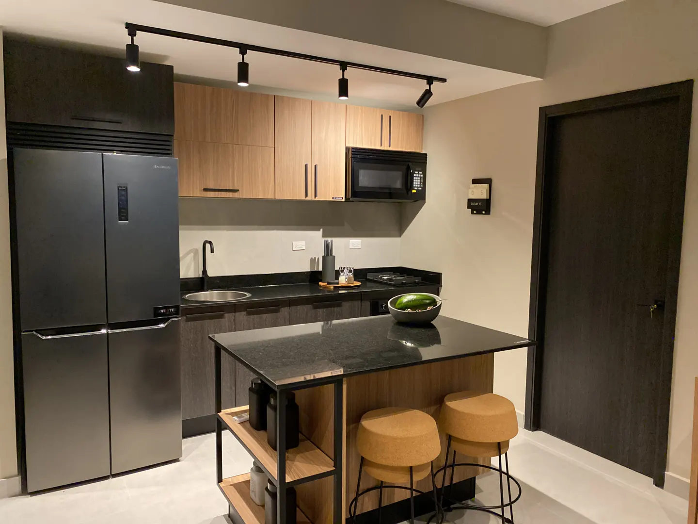 Modern kitchen with light wood cabinets, black countertops, stainless steel fridge, island with two stools, and track lights.