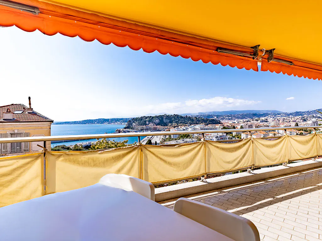 Balcony view with yellow awning, white lounge chairs, and a panoramic view of the city and ocean on a sunny day.