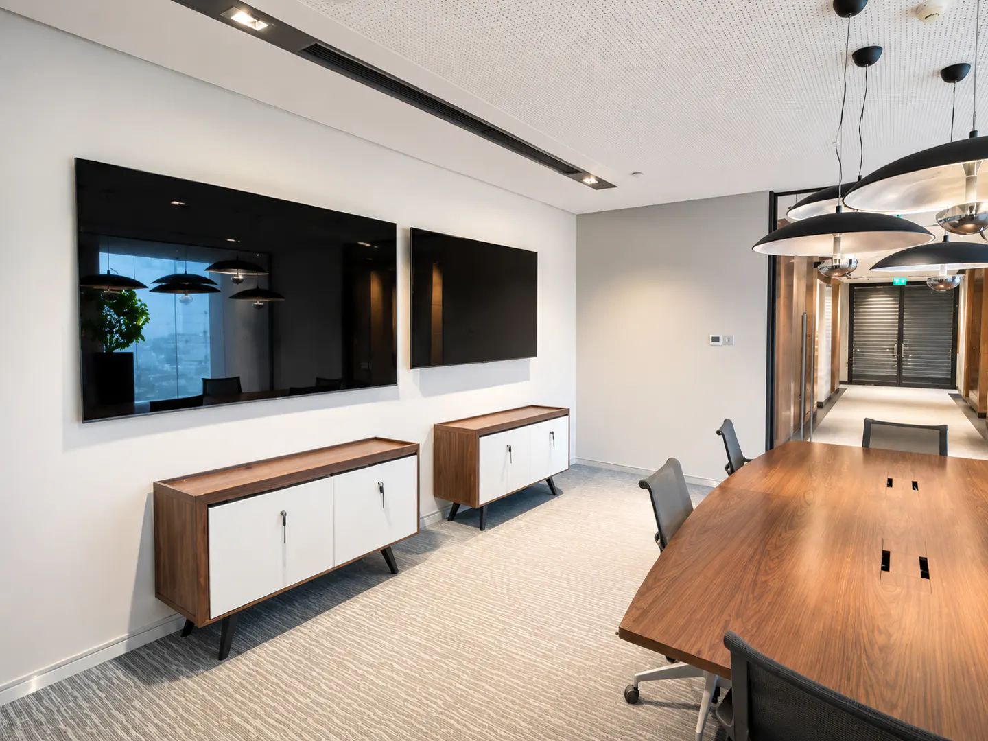 Modern office meeting room with a long wooden table, two wall-mounted TVs, and white cabinets on a grey patterned carpet.
