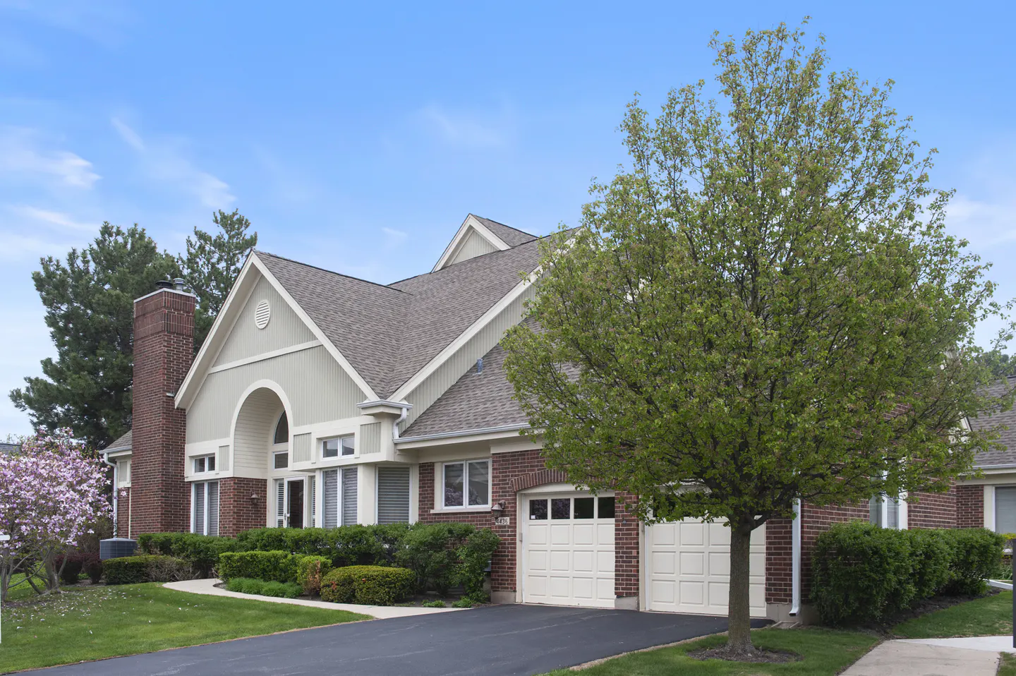 Brick house with a gray roof, white garage doors, and green lawn under a blue sky. A large green tree is in the foreground.