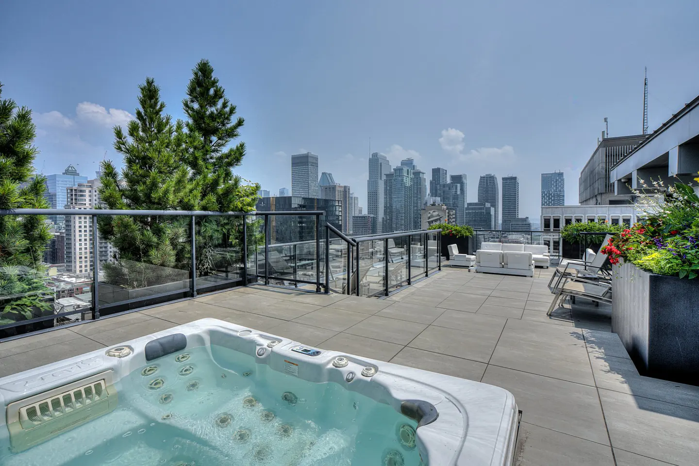 Rooftop terrace with a hot tub, lounge seating, and planters overlooking a city skyline under a clear blue sky.