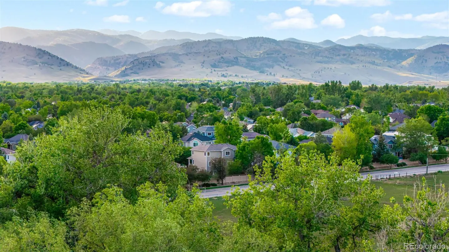 Scenic view of a neighborhood with lush green trees, houses, and mountains in the background under a blue sky.