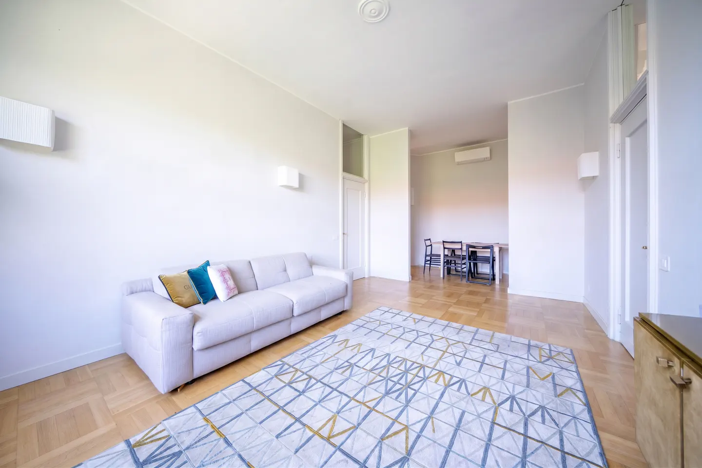 Bright living room with a white sofa, colorful pillows, and a geometric rug on a wood floor. A dining table is visible in the background.